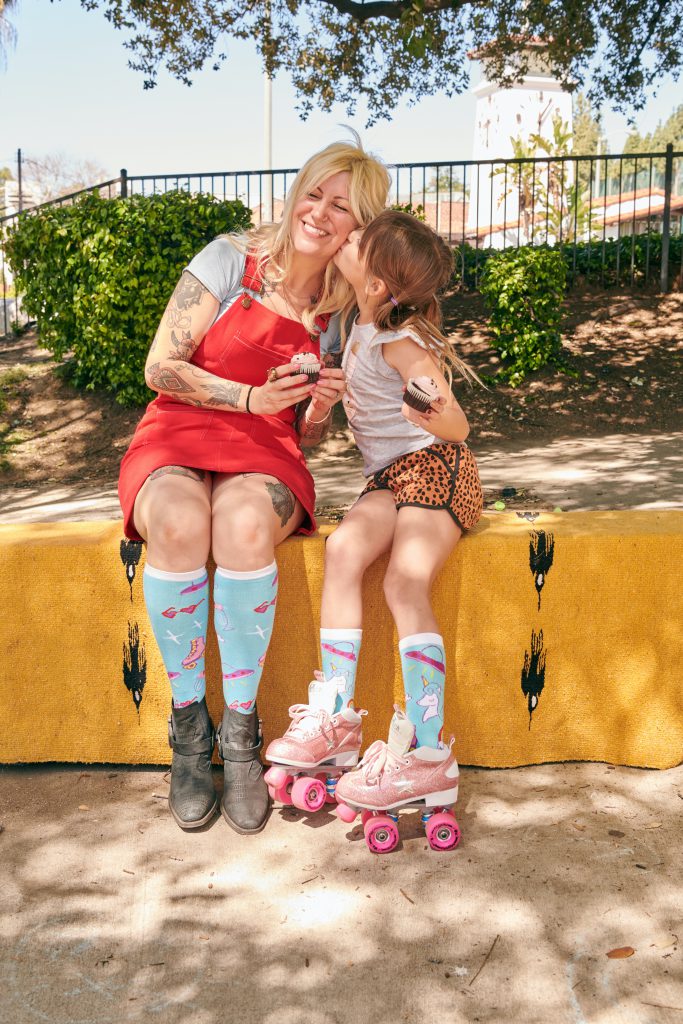 A mother and daughter sit outside. The daughter is kissing the happy mother's cheek. They're both wearing matching socks and eating cupcakes together. The little girl is in roller skates. 