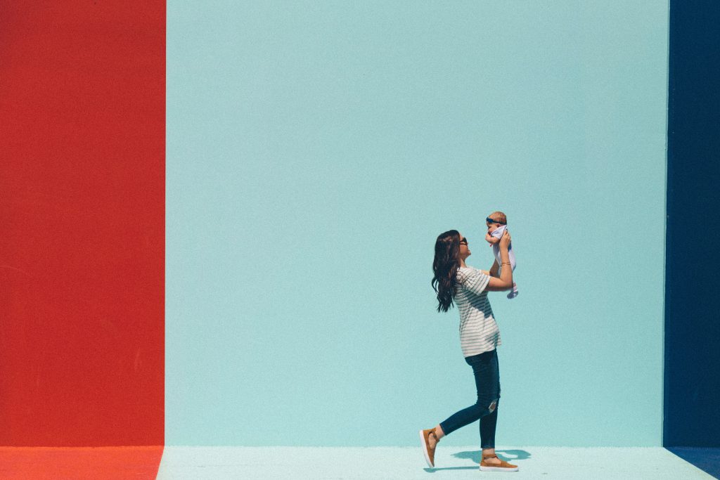 Mother holding baby while walking in front of a colorful wall.